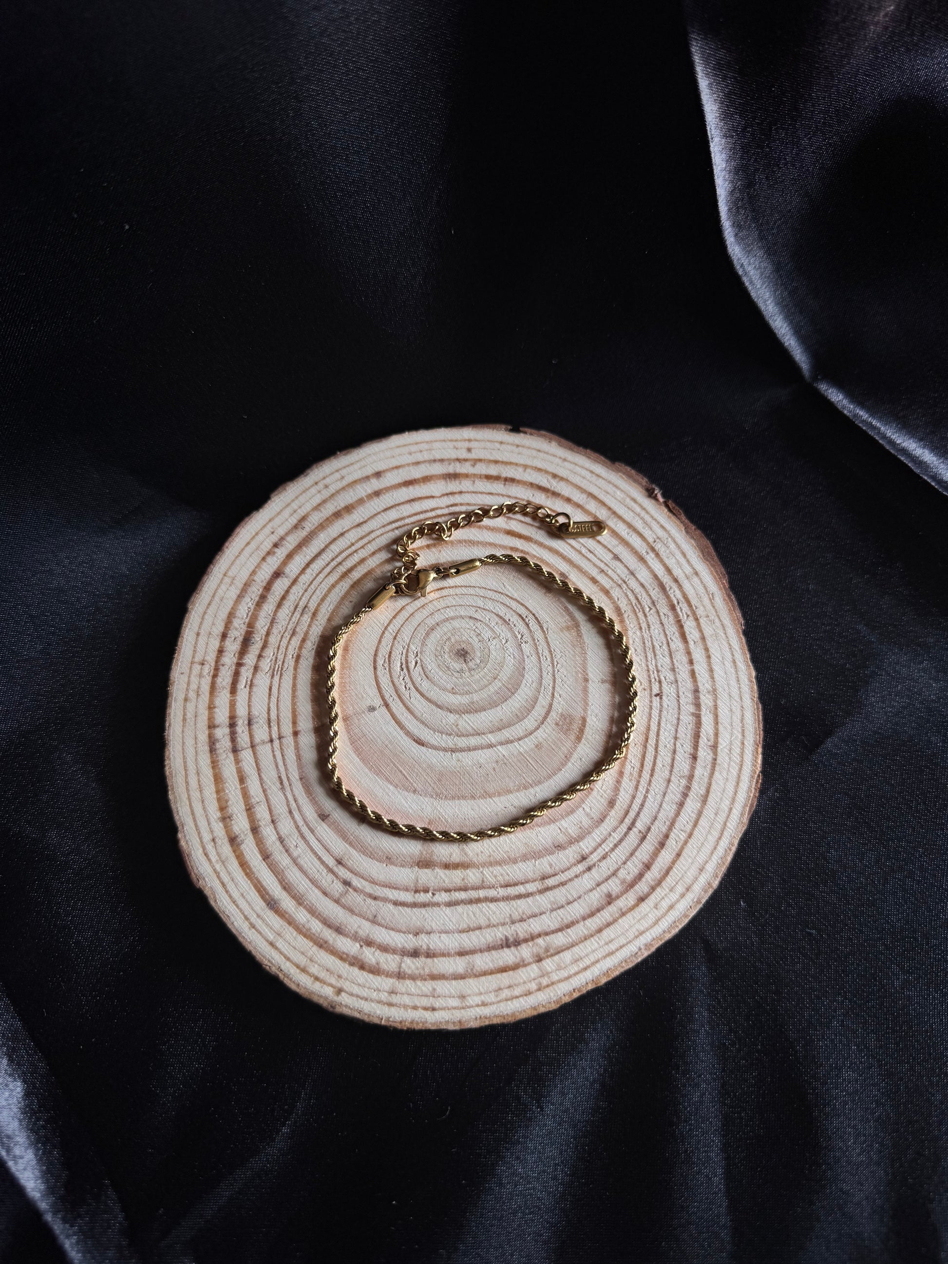 Gold rope anklet on a wooden disc with concentric circles against a dark fabric background