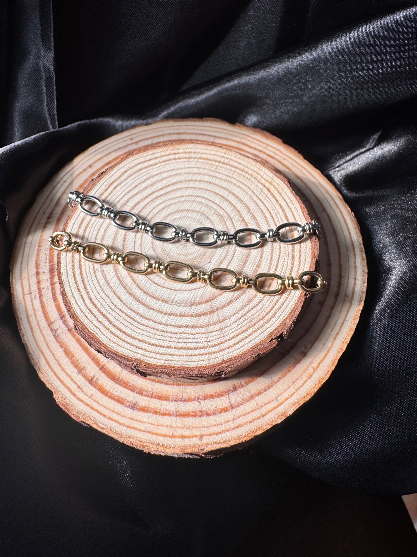 Pair of silver and gold anklets on a wooden surface with black satin fabric background