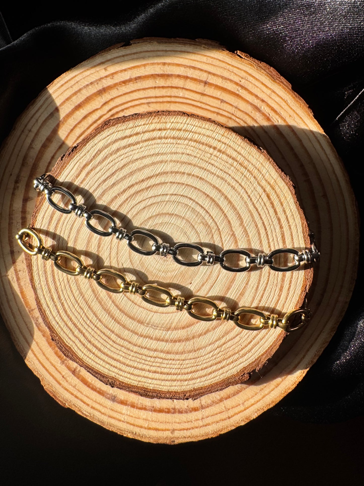 Two anklets, one silver and one gold, on a wooden surface with a black background.