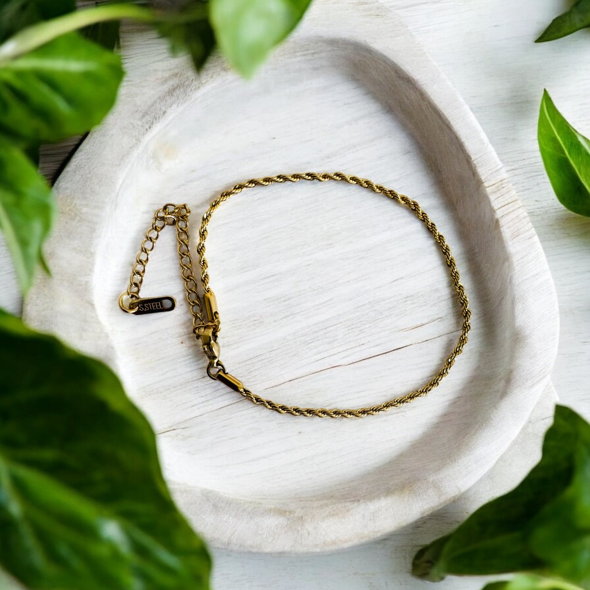 Gold rope anklet on a white stone surface with green leaves around