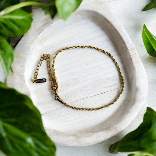 Gold rope anklet on a white stone surface with green leaves around