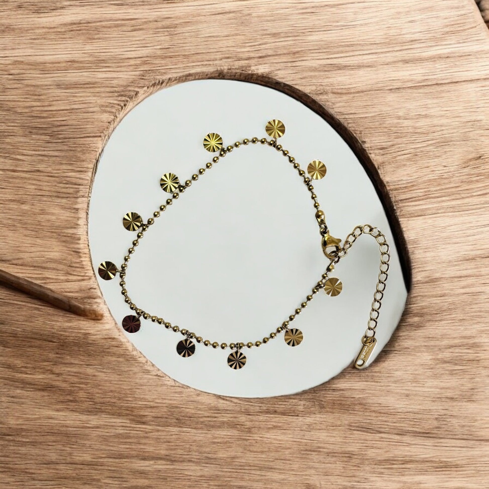 Gold anklet with disc elements on a white circular stand against a wooden background