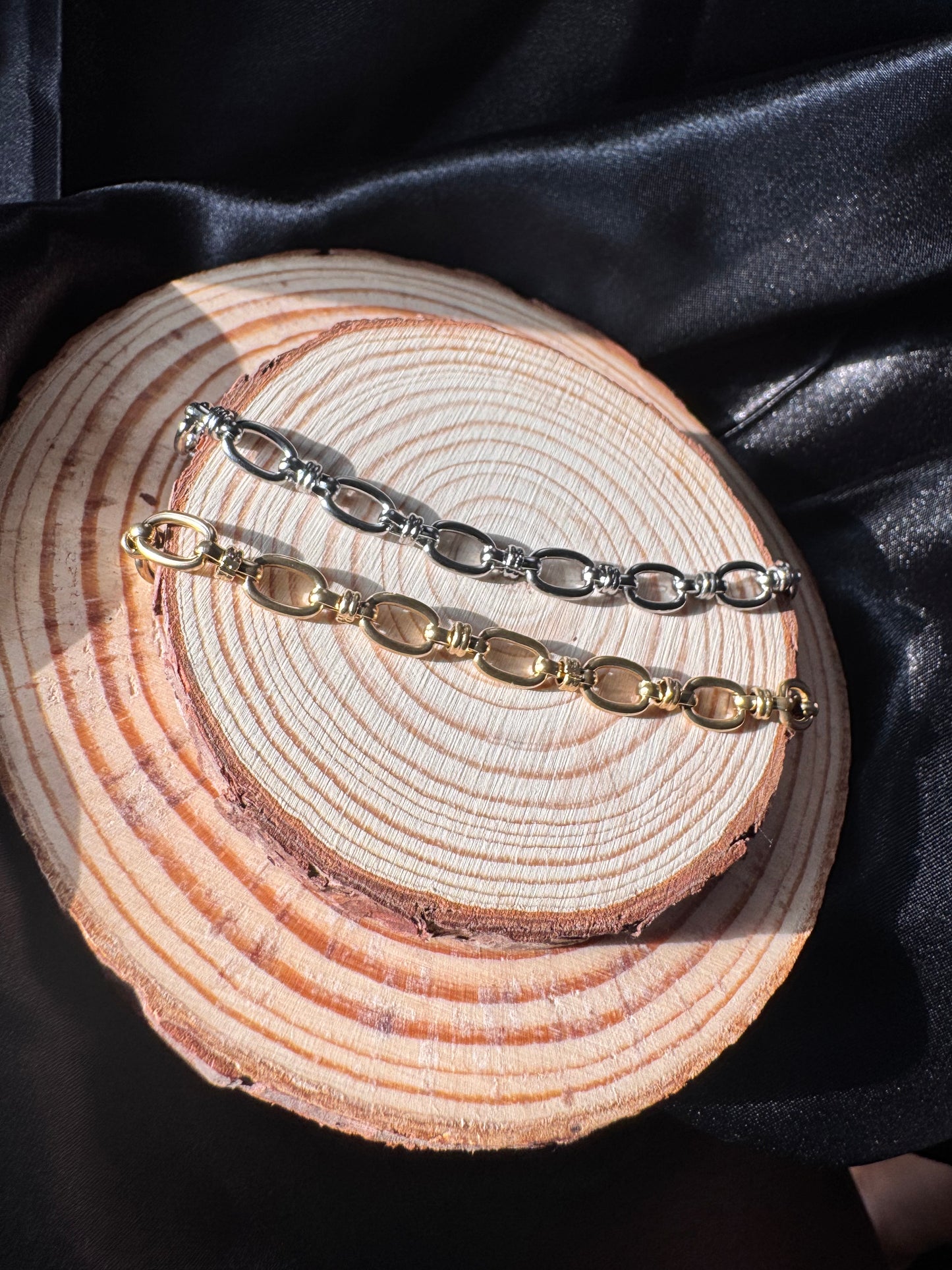 Two anklets, one silver and one gold, on a wooden surface with a dark fabric background.
